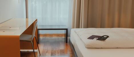 A hotel room with a wooden floor, a desk with a chair, a bed with a pillow, and a window with curtains.