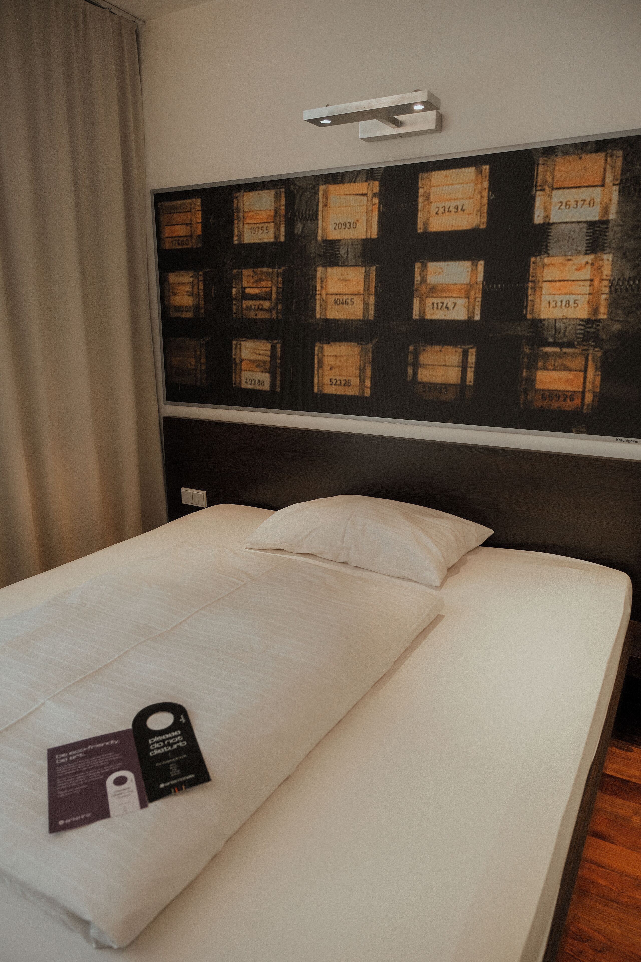 Habitación de hotel con una cama bien tendida, un cabecero con arte mural de cajas y una etiqueta en la cama.
