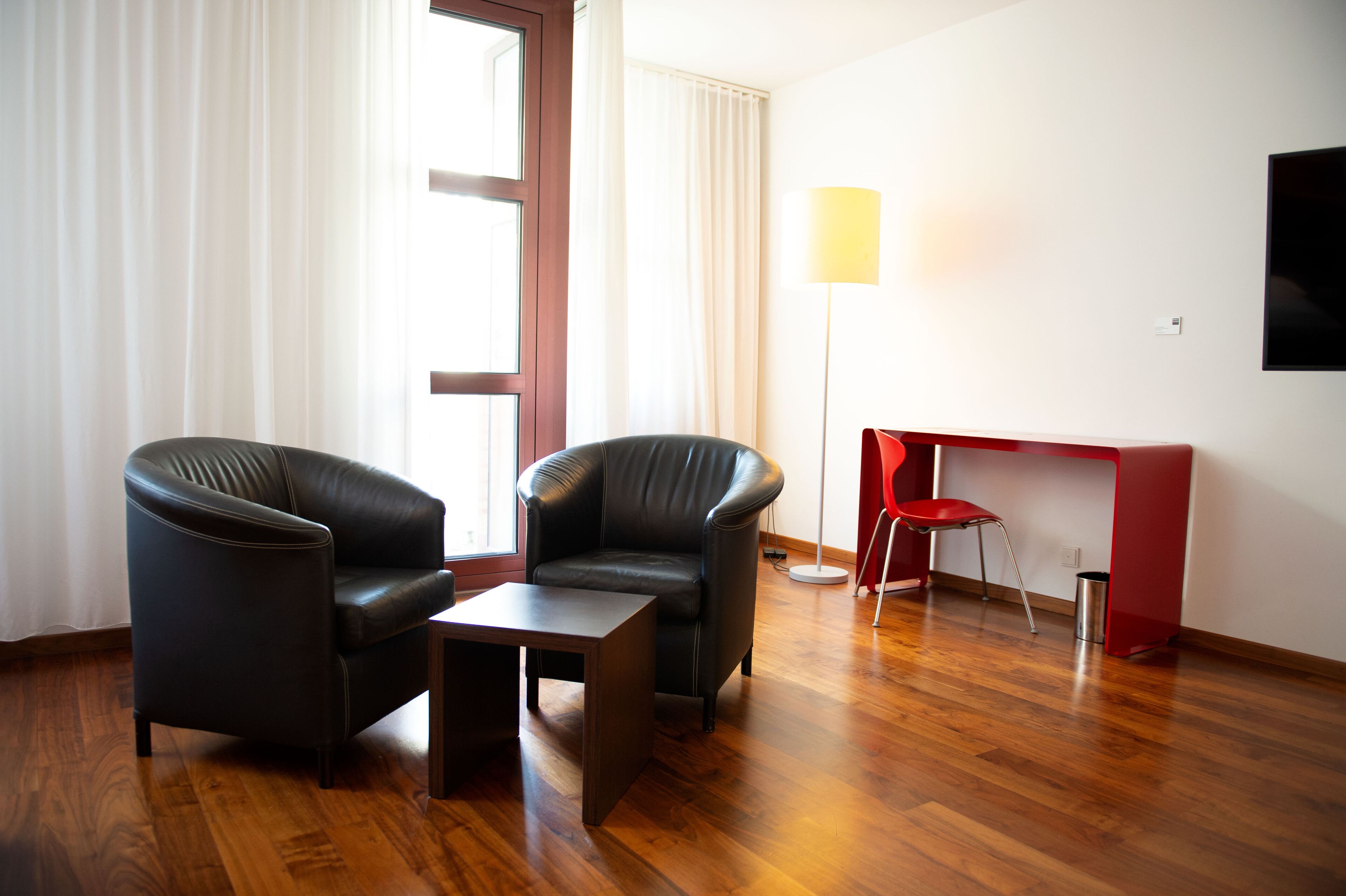 A room with two black leather chairs, a small table, a red chair, a red desk, and a floor lamp.