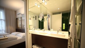 A hotel bathroom with two sinks, a large mirror, and a bed with white linens. - arte Hotel Linz (Linz)