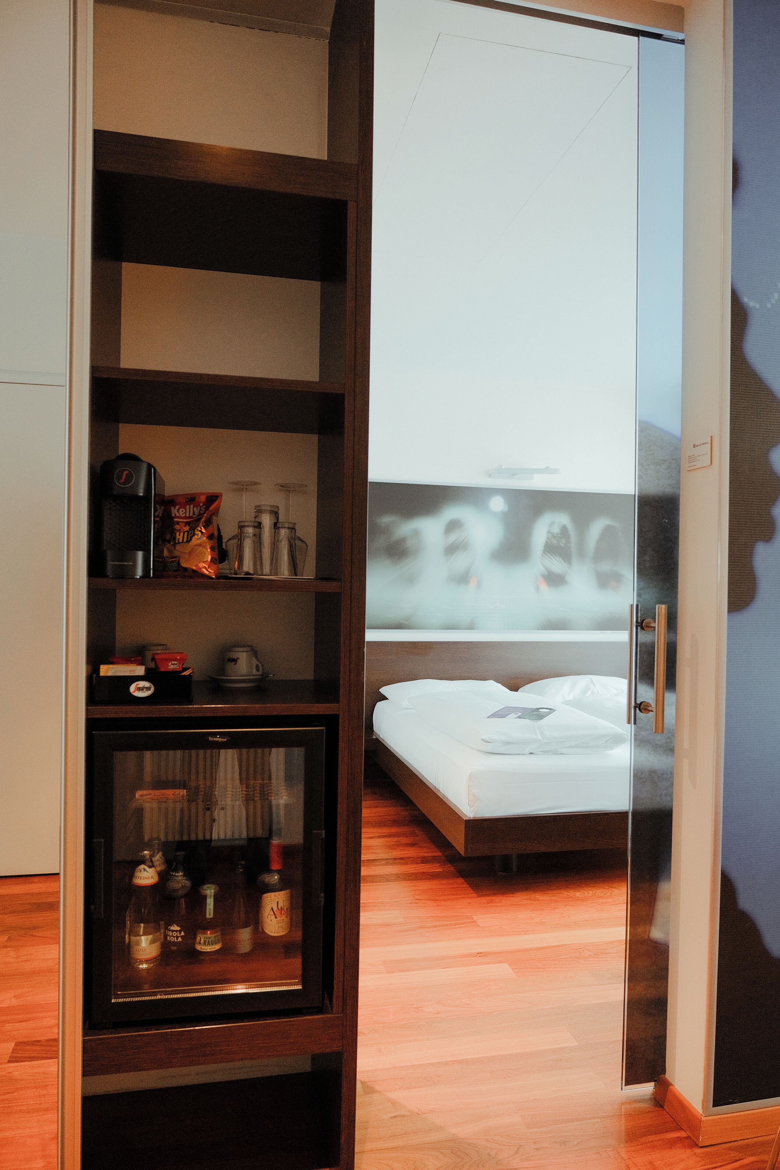 A hotel room with a glass door leading to a balcony, a bed, a minibar, and a shelf with various items.