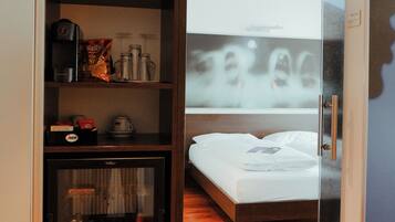 A hotel room with a glass door leading to a balcony, a bed, a minibar, and a shelf with various items.