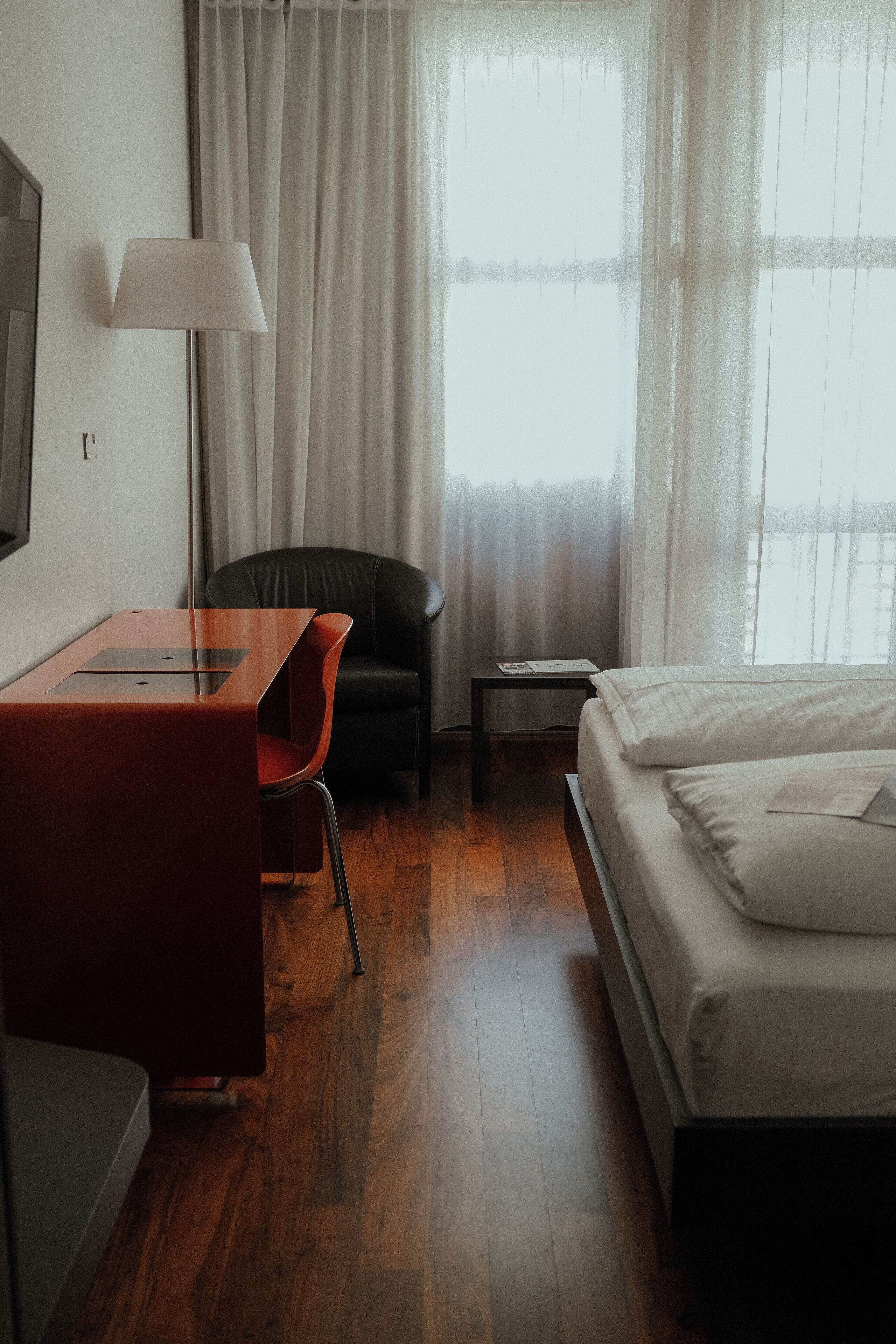 Ein Hotelzimmer mit Holzboden, einem Bett mit weißer Bettwäsche, einem Schreibtisch mit einem roten Stuhl, einem schwarzen Stuhl, einer Lampe und einem Nachttisch mit einem Buch.