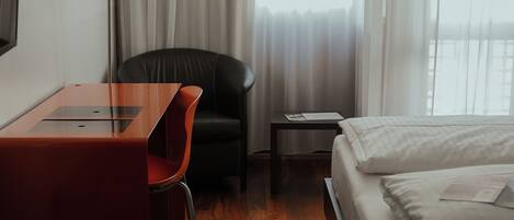 A hotel room with a wooden floor, a bed with white bedding, a desk with a red chair, a black chair, a lamp, and a nightstand with a book.