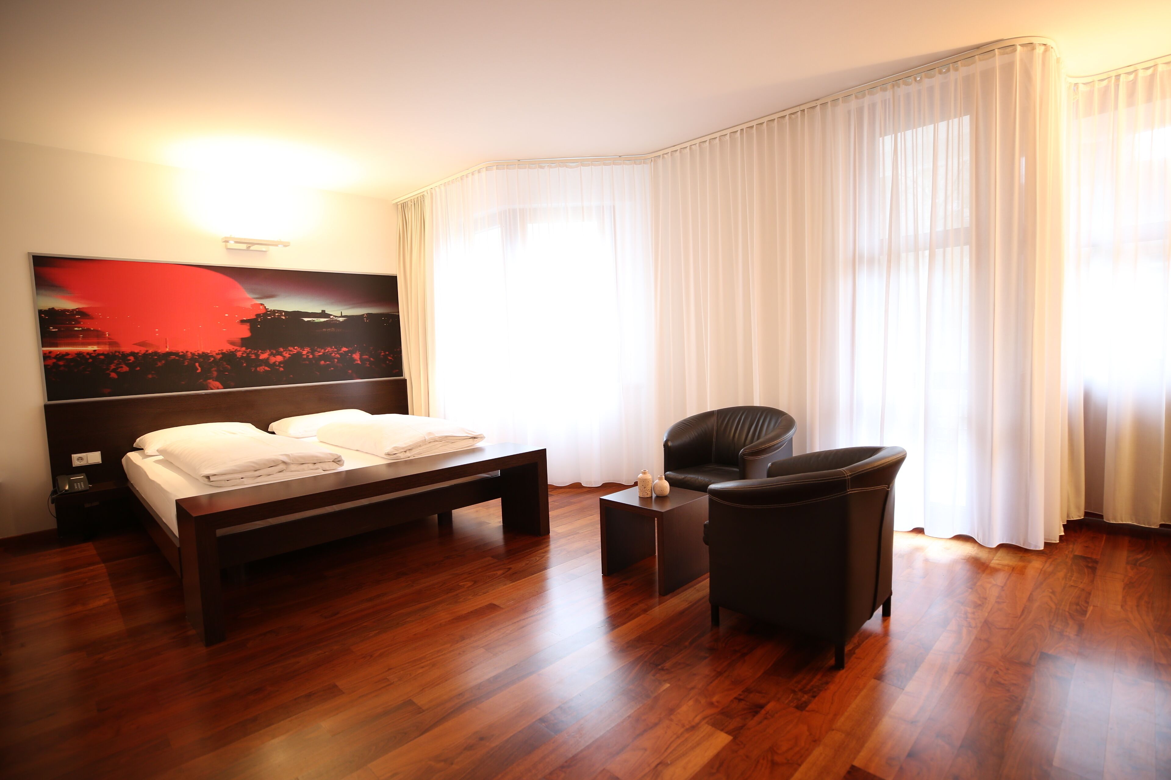 Een moderne hotelkamer met een houten vloer, een groot bed, een zwarte fauteuil, een klein tafeltje en een schilderij aan de muur.