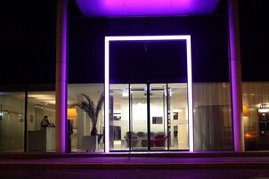 Una entrada de hotel moderna con fachada de vidrio, un mostrador de recepción y un área de descanso visible en el interior.