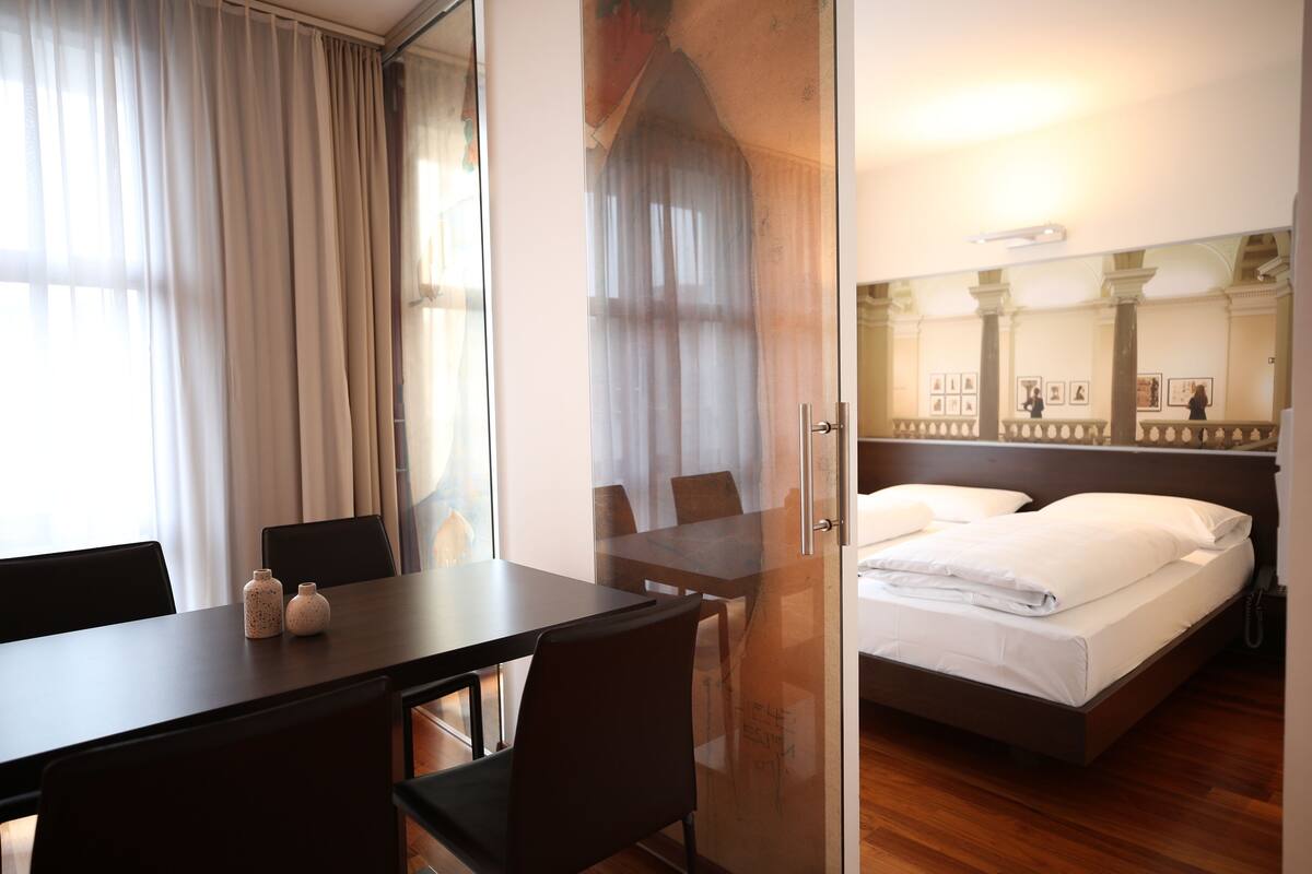 A hotel room with a bed, a desk, chairs, and a glass partition.