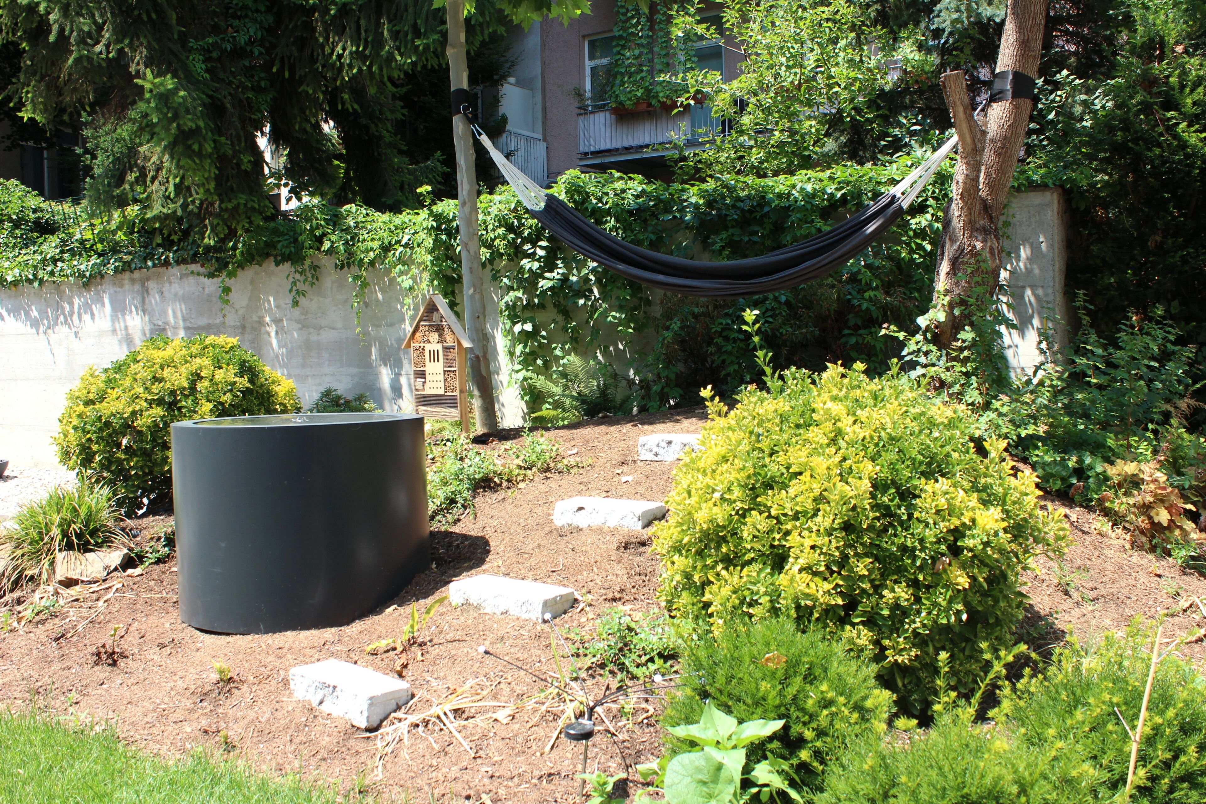 Un jardin avec un hamac, une nichoir et un grand pot de fleurs.