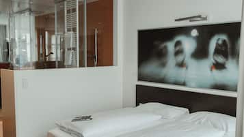 A modern hotel room with a large bed, a seating area, a large mirror, and a wall-mounted TV.