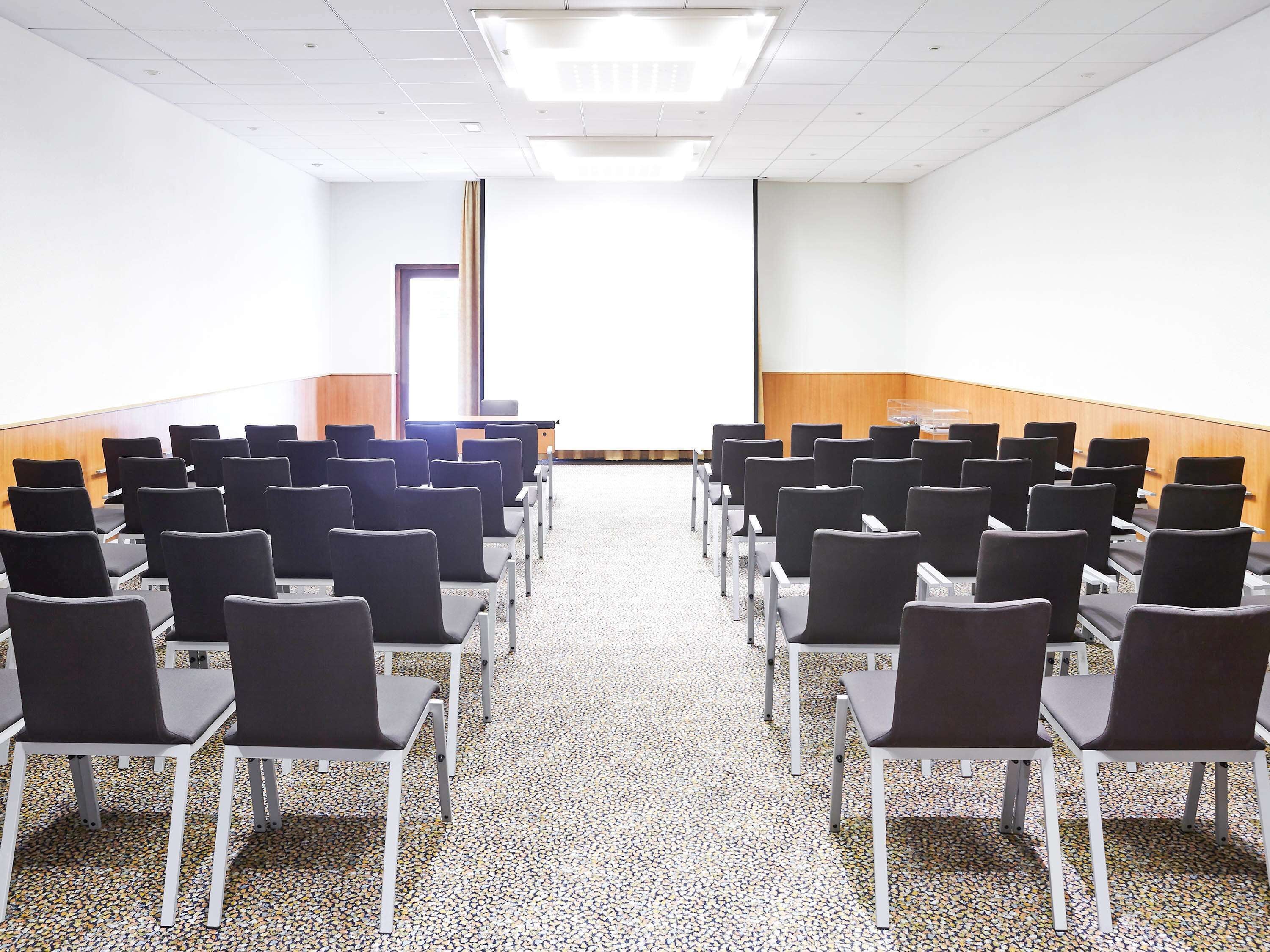 Une salle de conférence avec des rangées de chaises, un écran de projection et un sol recouvert de moquette.