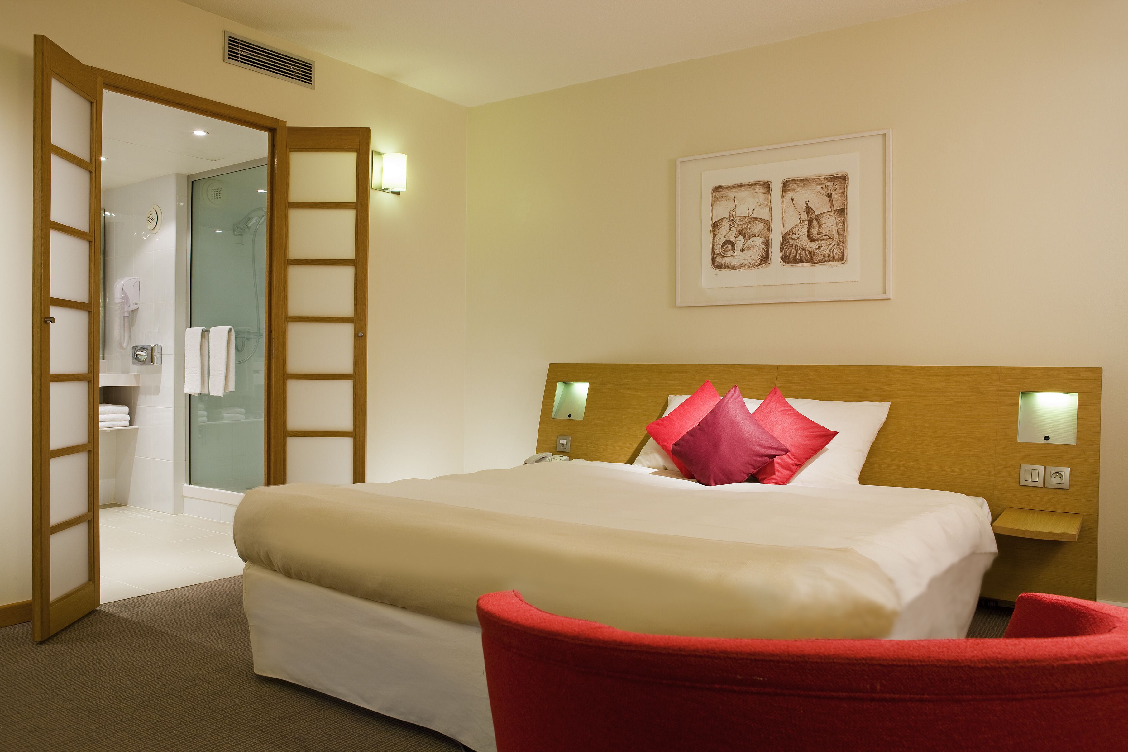A hotel room with a large bed, a red chair, a wooden headboard, a framed picture on the wall, and a bathroom visible through an open door.