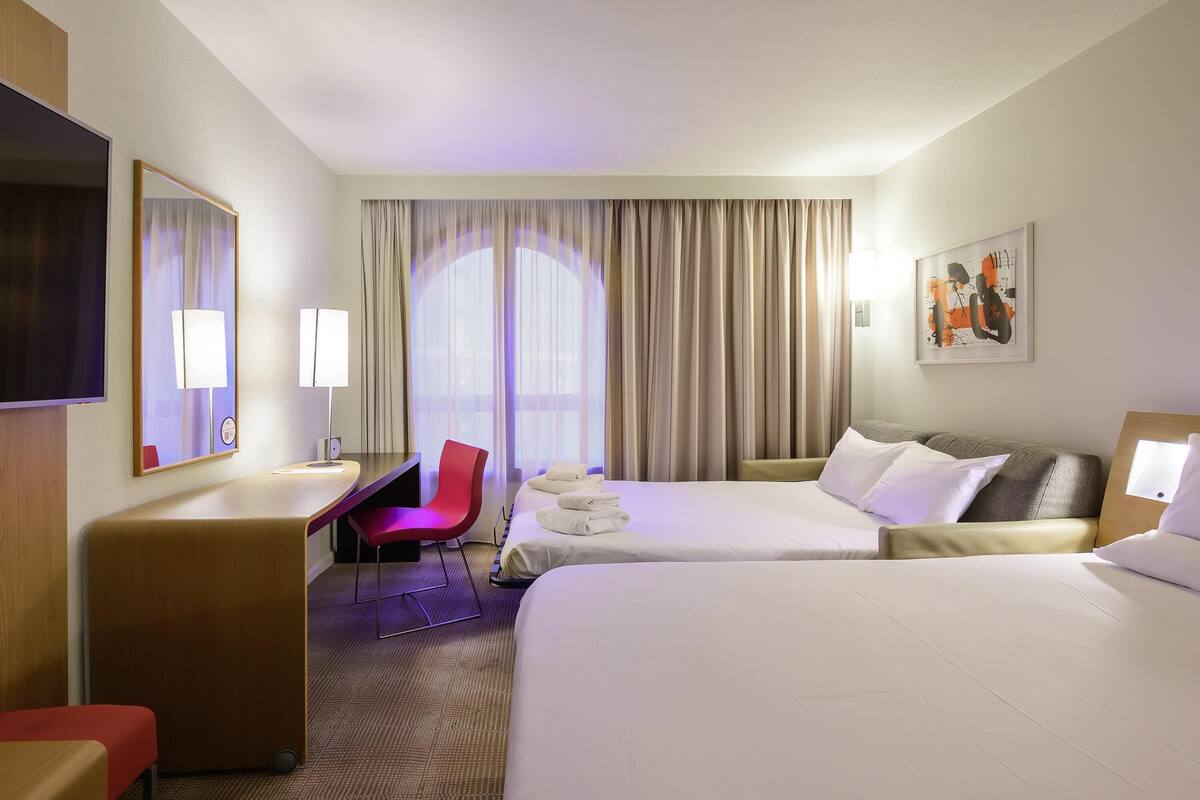A hotel room with two beds, a desk, a chair, a TV, and a large window with curtains.