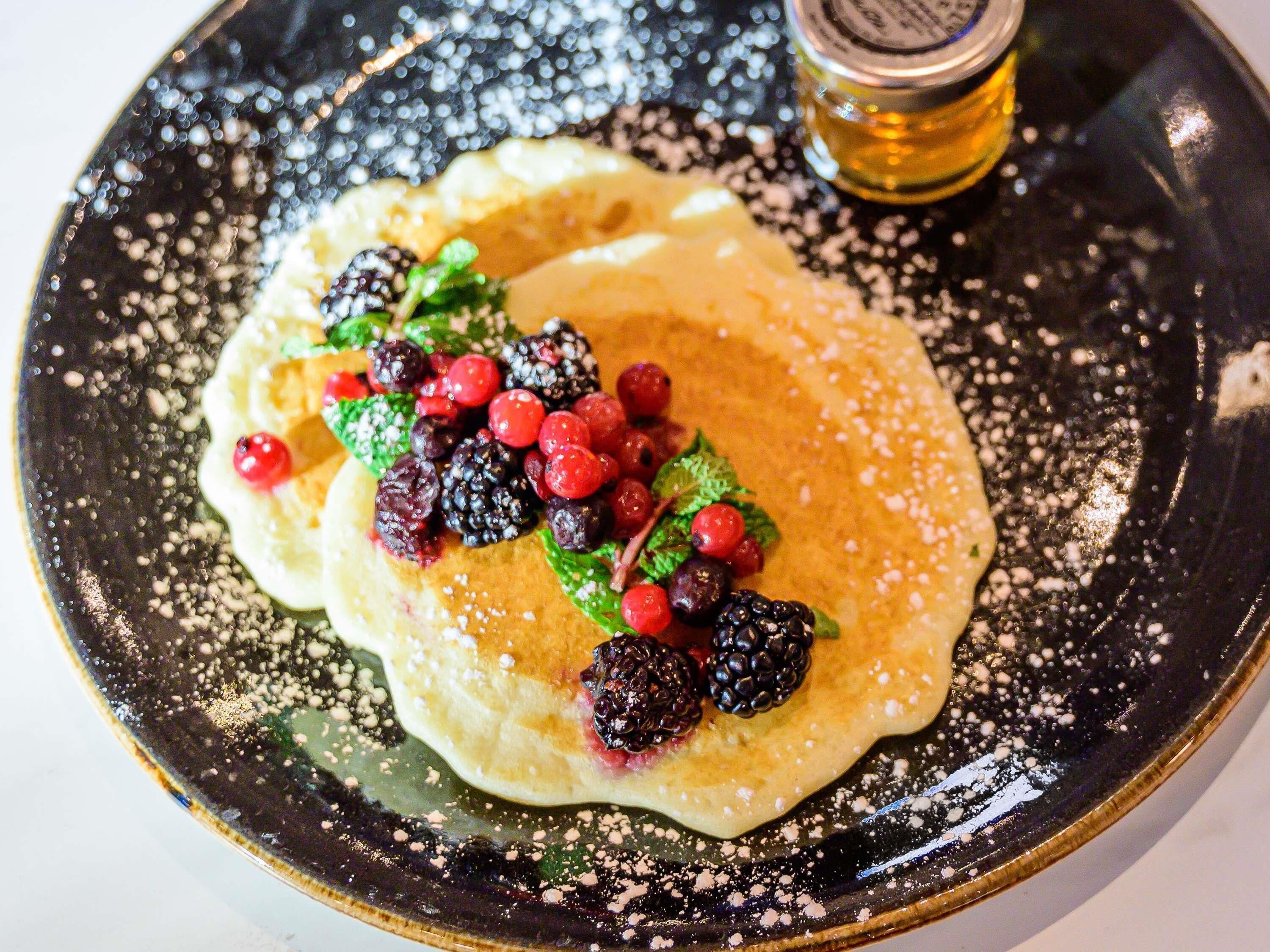 A plate with pancakes topped with berries and mint, dusted with powdered sugar, and a bottle of syrup.