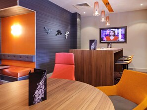 Ein modernes Hotelzimmer mit einer Holztresenrezeption, einem an der Wand montierten Fernseher, einer Sitzecke mit orangefarbenen und roten Stühlen sowie einem großen Fenster.
