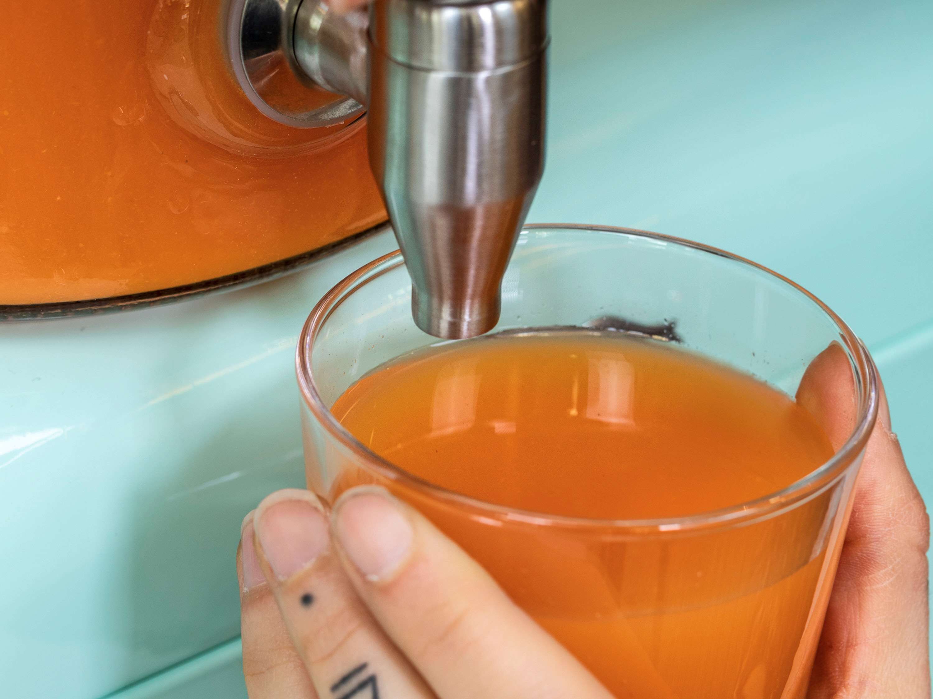 A hand holding a glass filled with a liquid, being filled from a metallic dispenser.