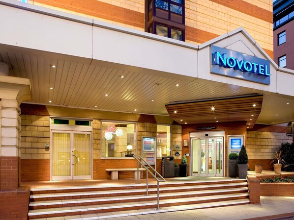The exterior of a hotel with a prominent 'Novotel' sign, a set of stairs leading to the entrance, and a brick facade.