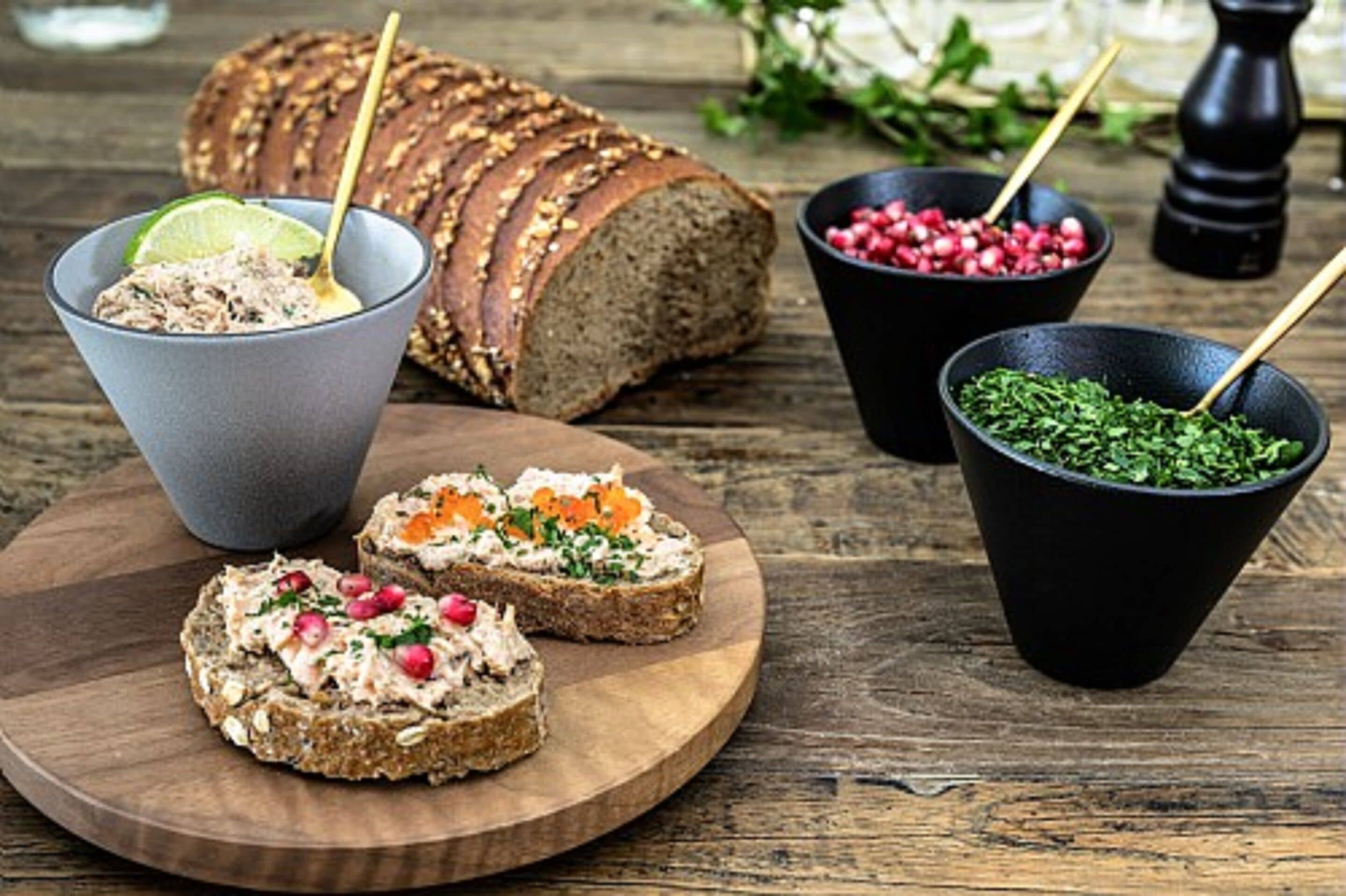 A wooden board with slices of whole grain bread, a bowl of spread with pomegranate seeds, and a bowl of green herbs.