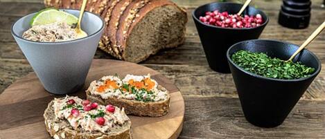 Une planche en bois sur laquelle sont disposées des tranches de pain complet, un bol contenant une pâte à tartiner aux graines de grenade et un bol d’herbes fraîches.