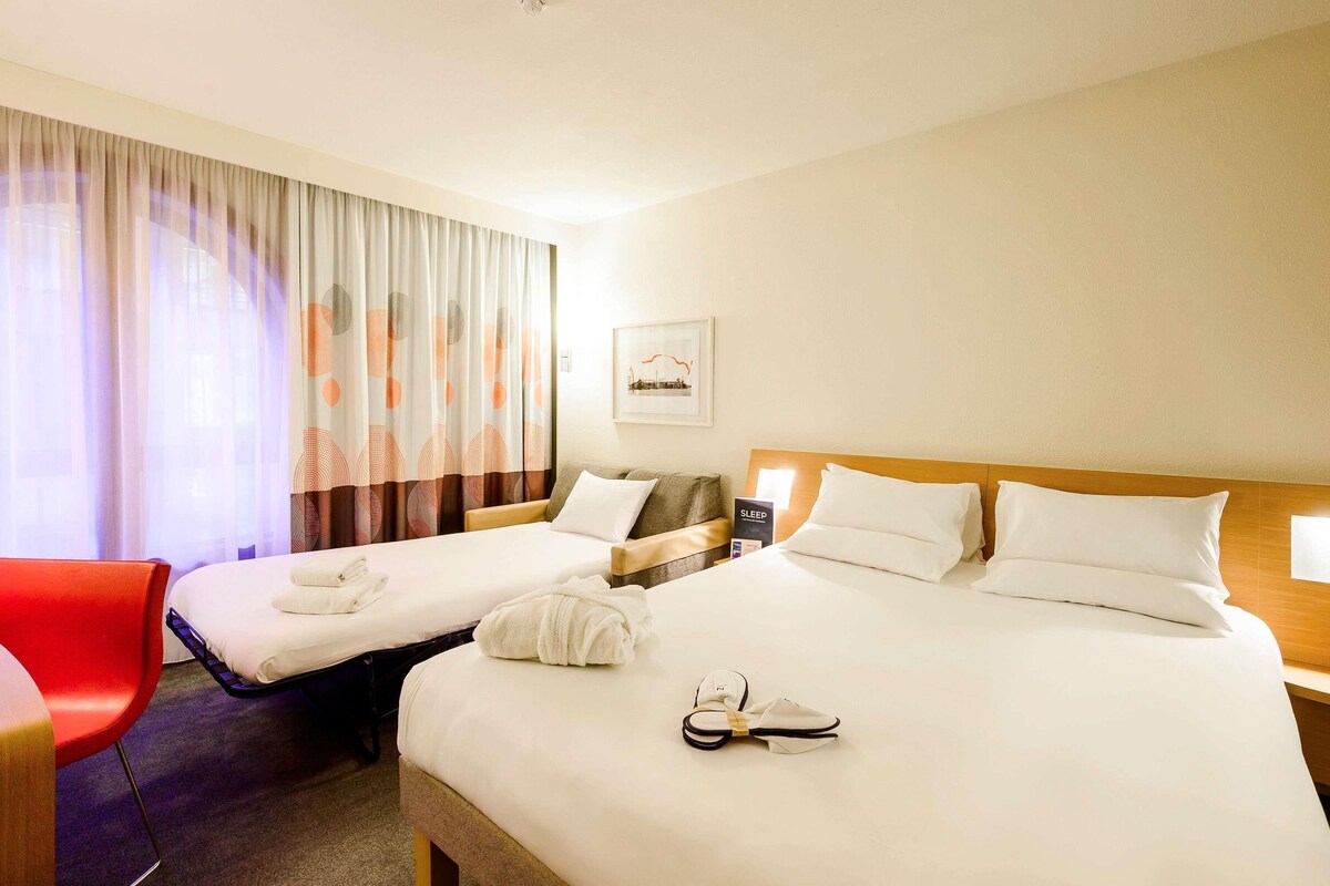 A hotel room with two beds, a red chair, a bedside table with a lamp, and a framed picture on the wall.