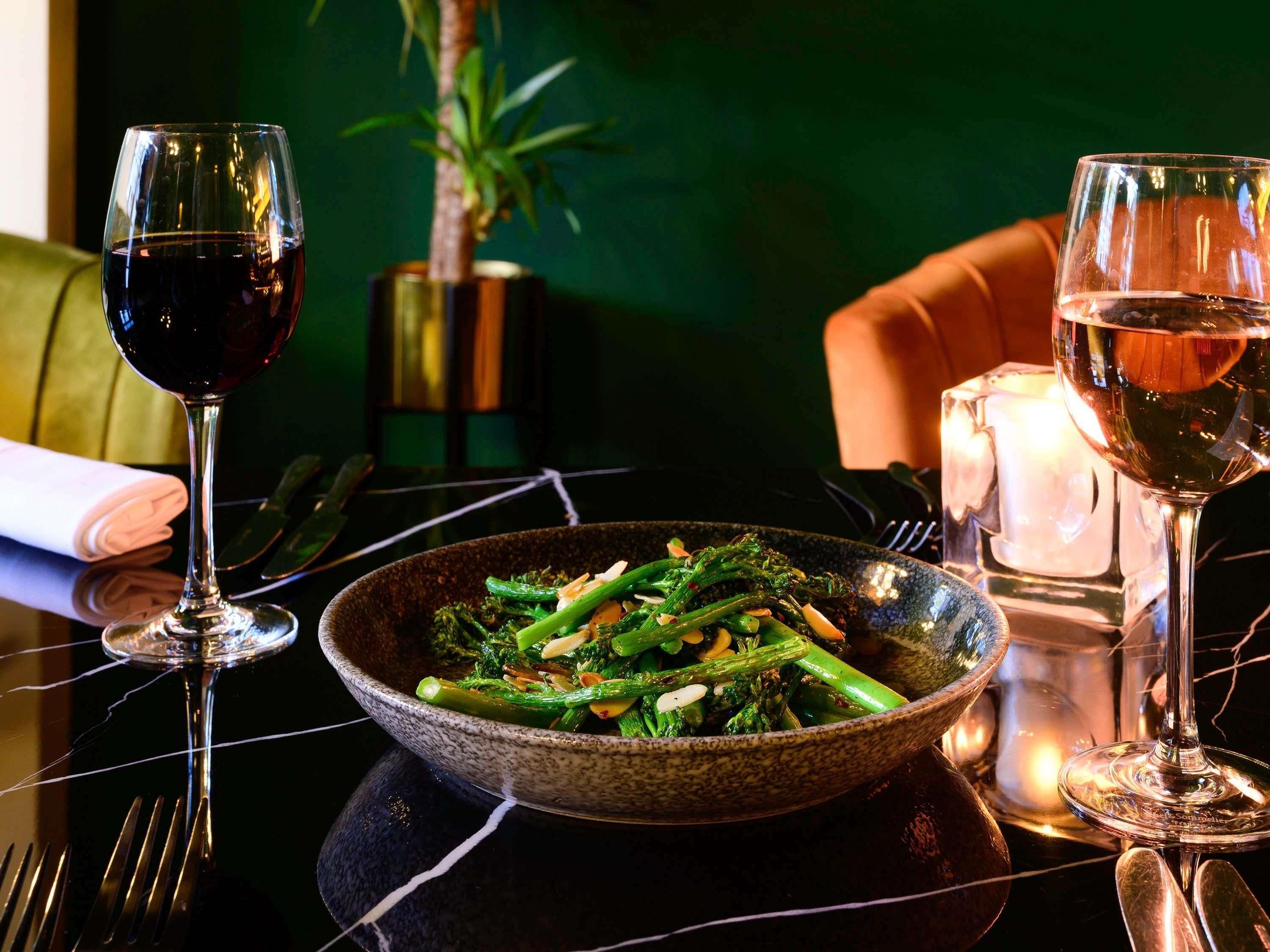 A dining table with a bowl of green vegetables, two wine glasses, and a candle.