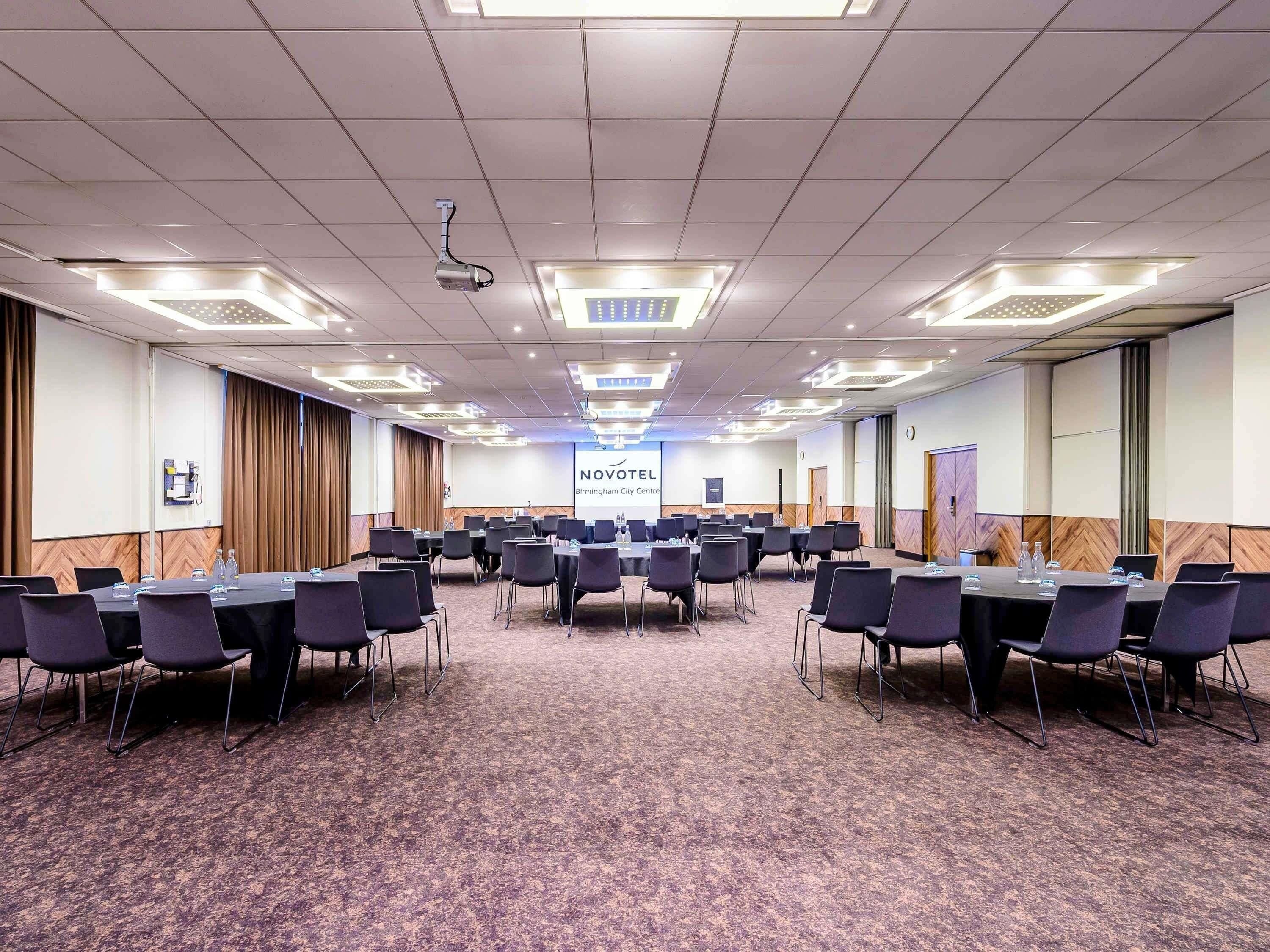 A conference room with multiple tables and chairs, a projector, and a screen displaying 'NOVOTEL Birmingham City Centre'.