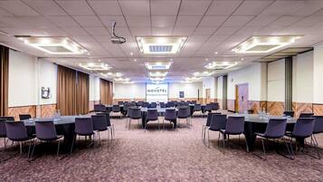 A conference room with multiple tables and chairs, a projector, and a screen displaying 'NOVOTEL Birmingham City Centre'.