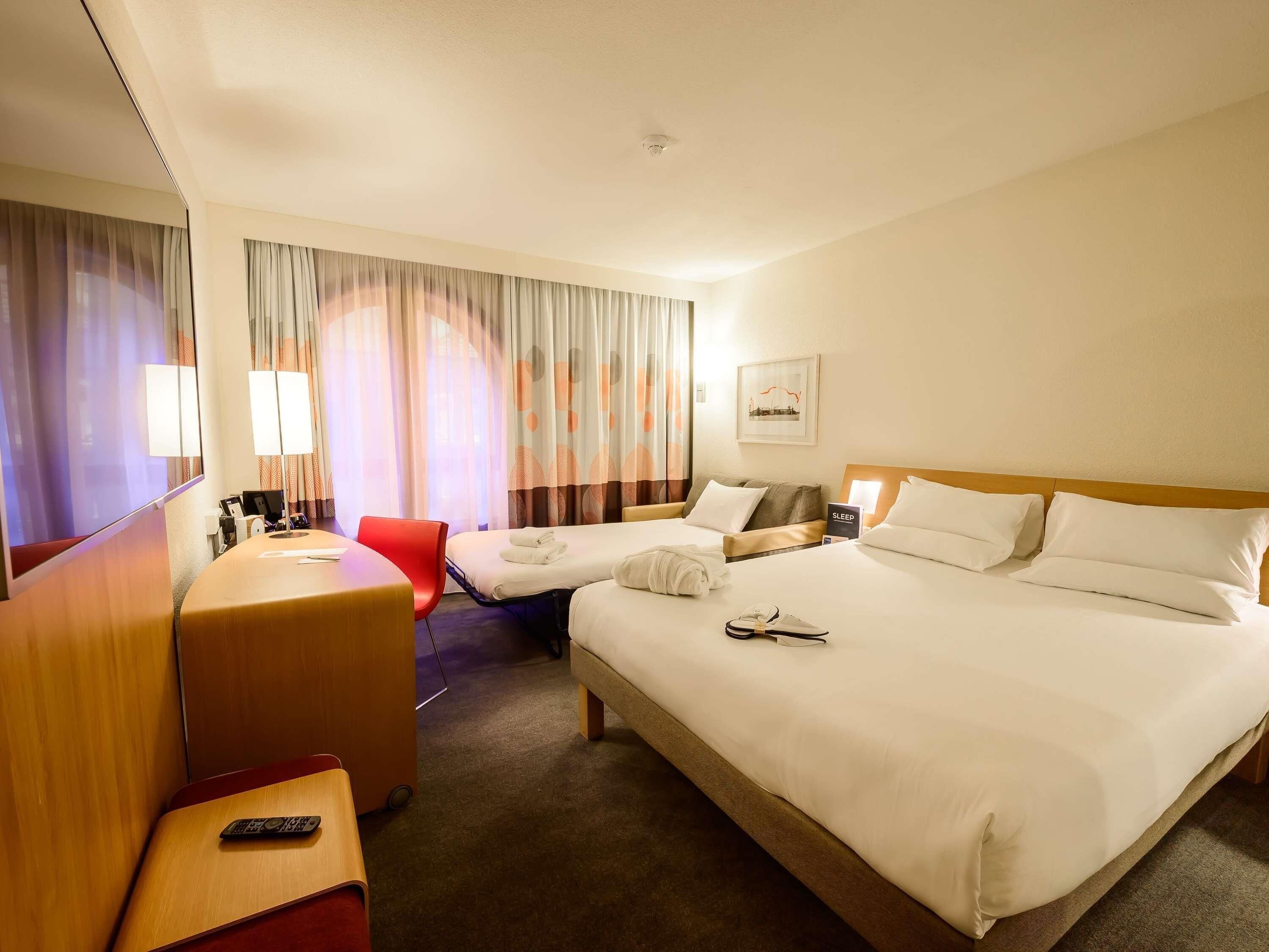 A hotel room with a large bed, a desk with a chair, a small table, and a TV.