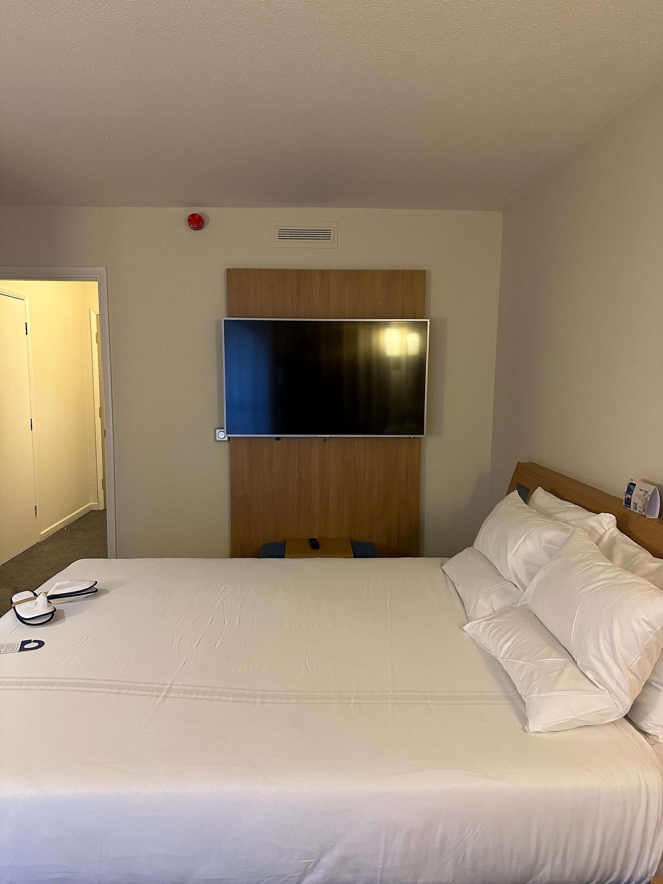 A hotel room with a bed, a television mounted on the wall, a door, and a small table with a remote control.
