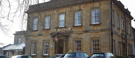 Edificio histórico identificado como “The Manor”, con un estilo arquitectónico clásico, múltiples ventanas y una entrada destacada. Varios coches aparcados en la parte delantera, lo que indica que es una propiedad pública o comercial.