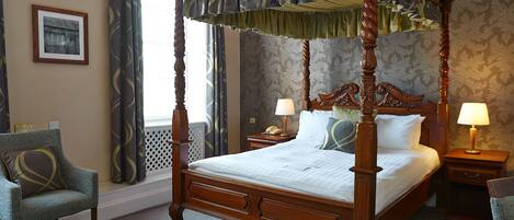 Uma cama com dossel e cortina, uma mesa de cabeceira com abajur, uma cadeira com almofada e um quadro pendurado na parede.