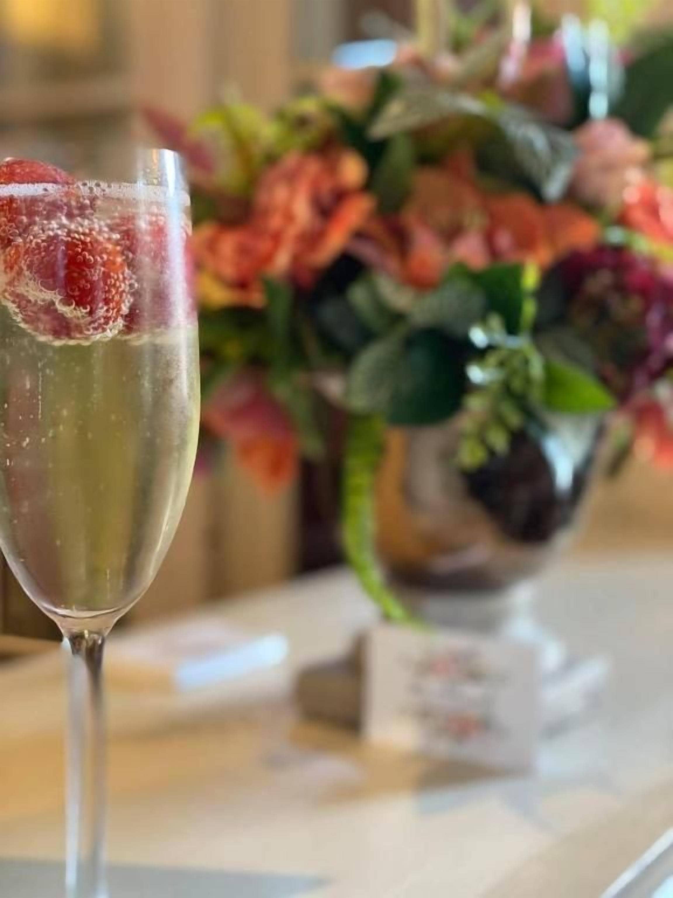 Un bicchiere di bevanda trasparente con una fragola, su un tavolo con un vaso di fiori sullo sfondo sfocato.
