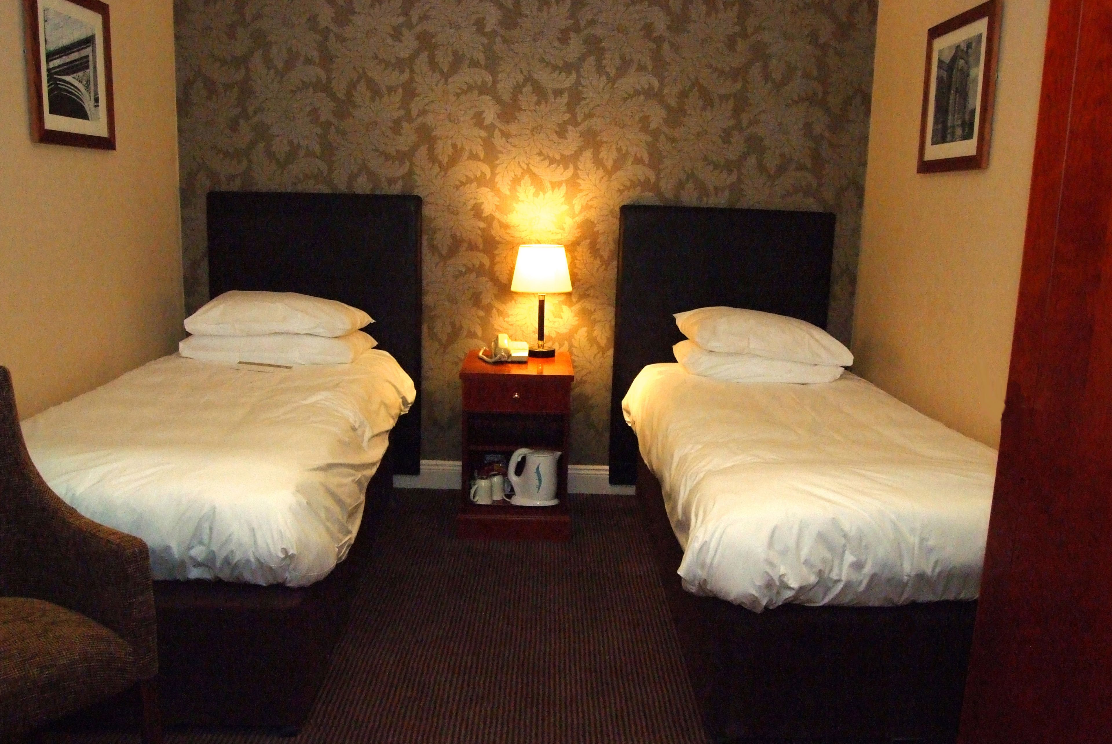 A hotel room with two single beds, a nightstand with a lamp, and a chair.