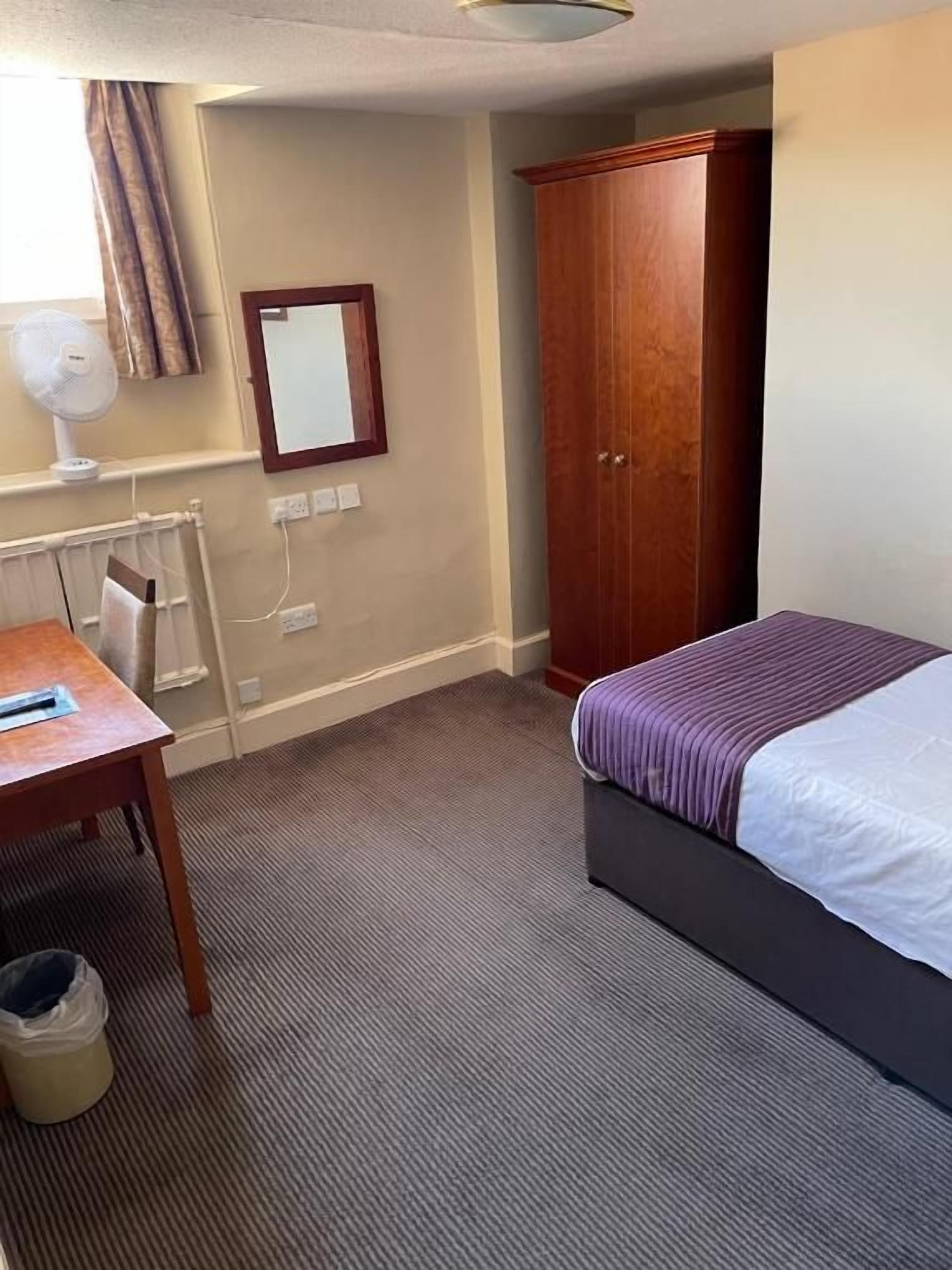 A hotel room with a bed, a desk, a wardrobe, a radiator, and a mirror.