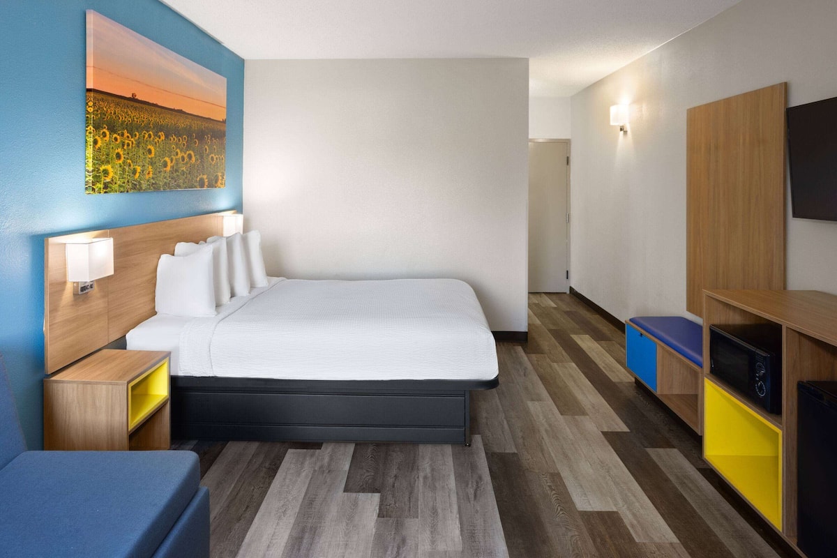 Habitación de hotel con cama, lámparas de noche, televisor, cabecera de madera y un cuadro en la pared.