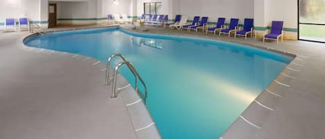 Una amplia piscina interior con agua cristalina azul, rodeada de sillas de descanso azules y blancas, y un suelo de baldosas.