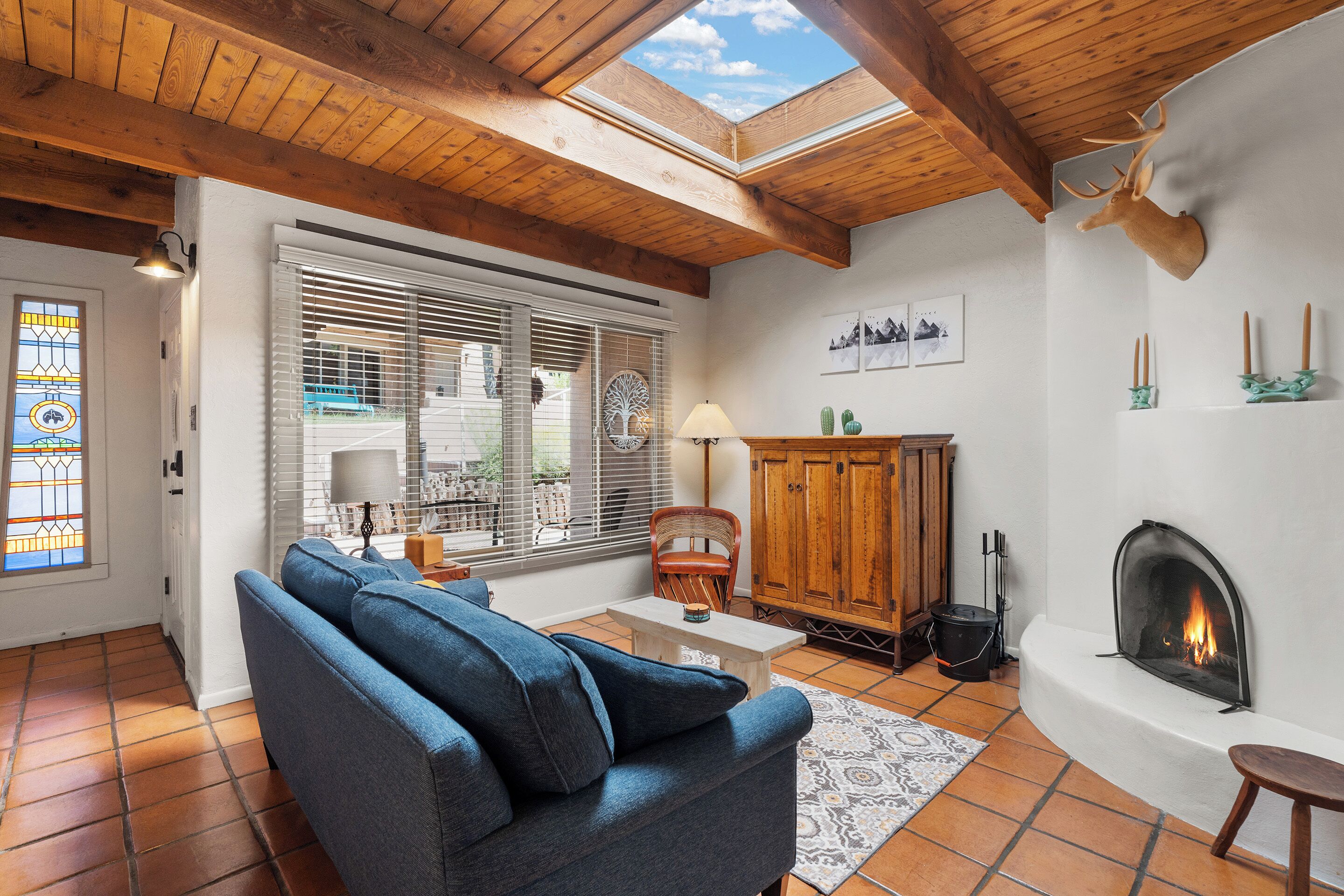 A cozy living room with a blue sofa, wooden ceiling beams, a fireplace, and a large window with blinds.