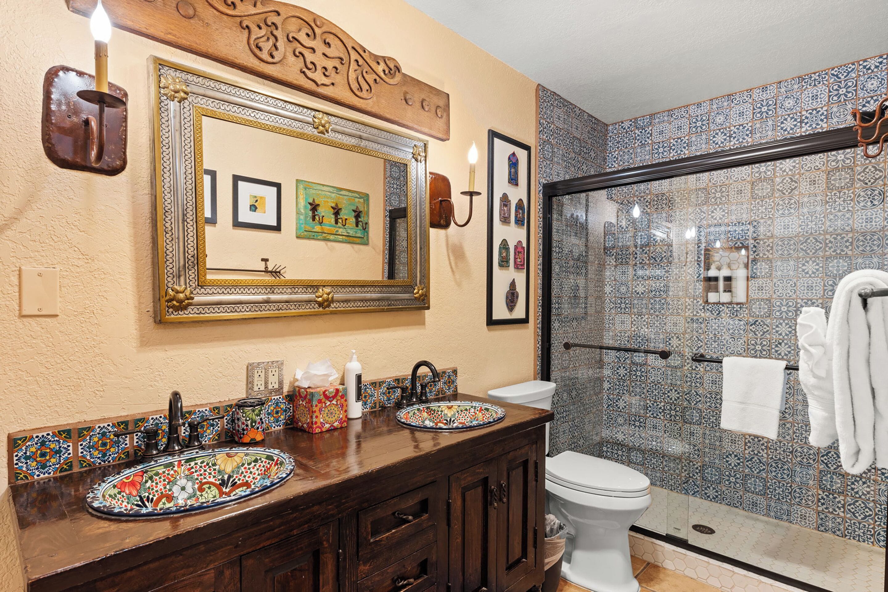 A bathroom with a large ornate mirror, two sinks with decorative tiles, a toilet, and a glass shower enclosure.