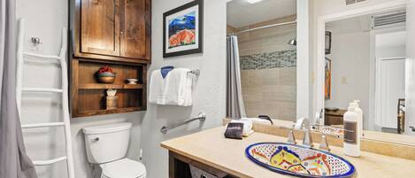 Un baño con inodoro, ducha, lavabo con un plato decorativo, espejo y armarios de madera.
