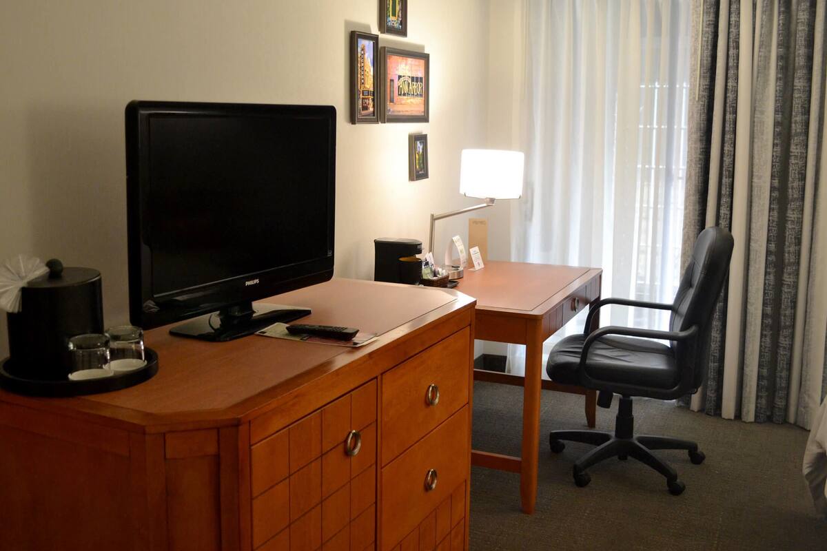 Une chambre d’hôtel avec un bureau en bois, une chaise de bureau noire, une télévision, une lampe et une machine à café.