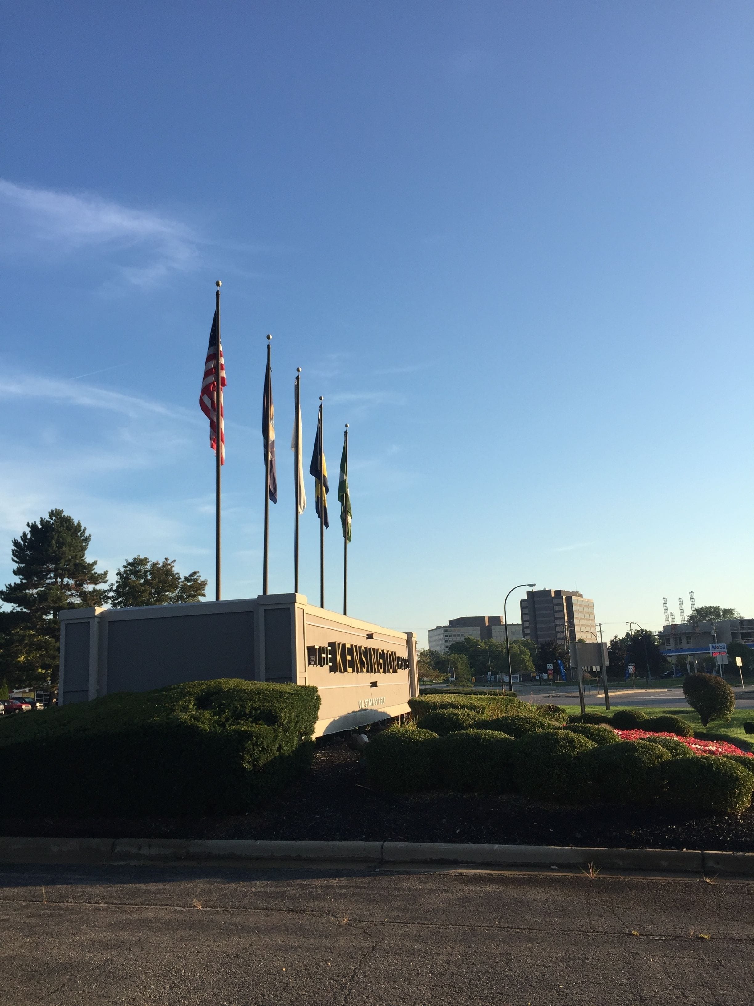 A sign with "The KENSINGTON" written on it, surrounded by flags, with a building and a Mobil gas station in the background.