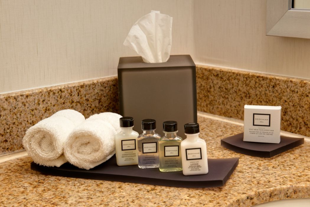 A hotel bathroom counter with a tissue box, rolled towels, and various toiletries including a conditioner, shampoo, and lotion.