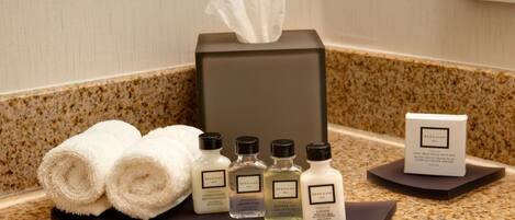 A hotel bathroom counter with a tissue box, rolled towels, and various toiletries including a conditioner, shampoo, and lotion.
