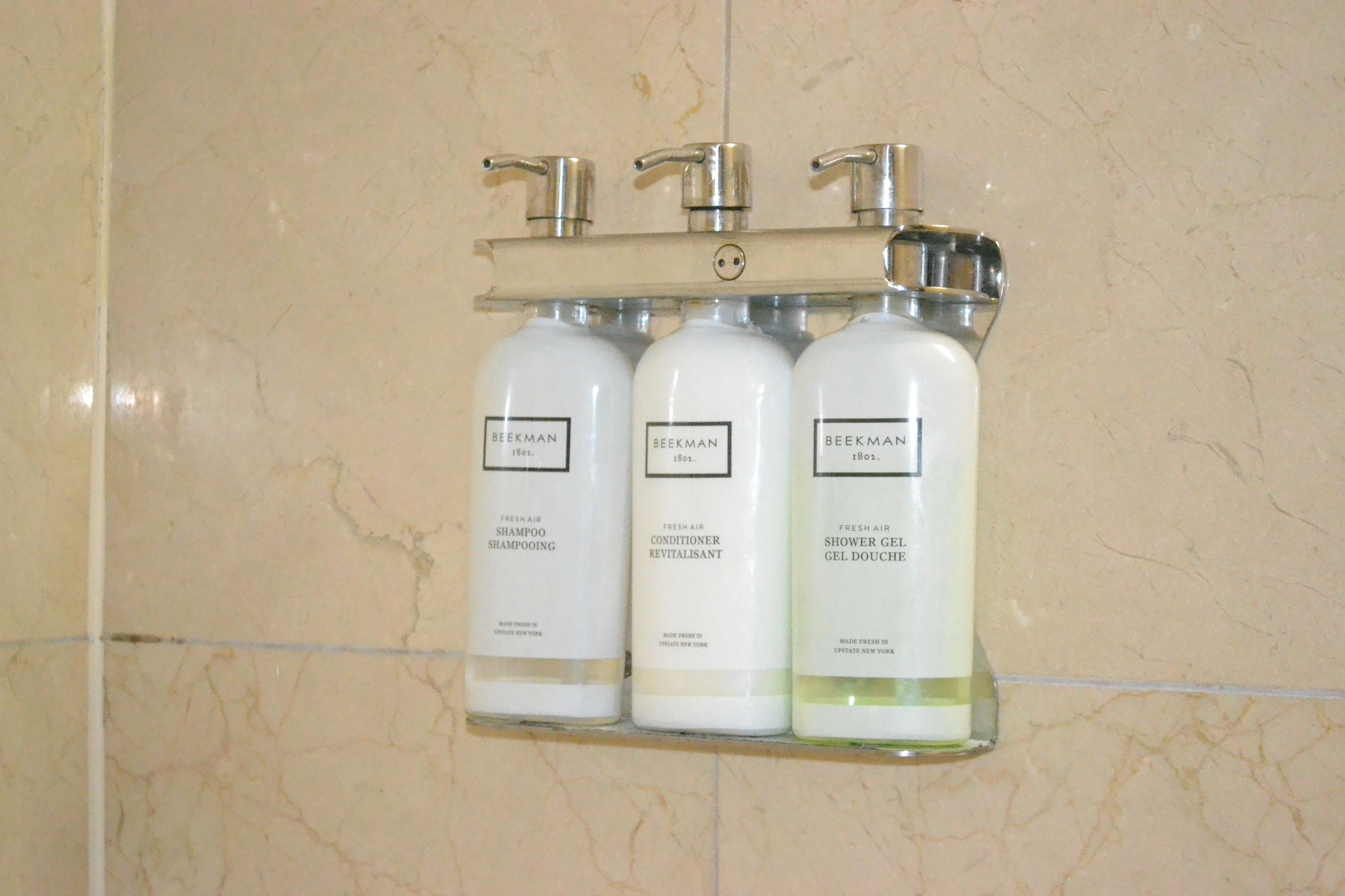 A bathroom with a marble wall and a shelf holding three bottles of Beekman products.