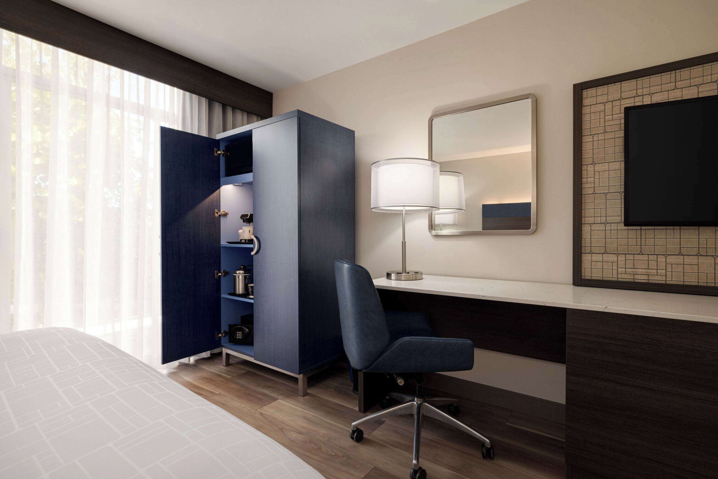 A hotel room with a bed, a desk with a chair, a wardrobe, a lamp, and a TV on the wall.