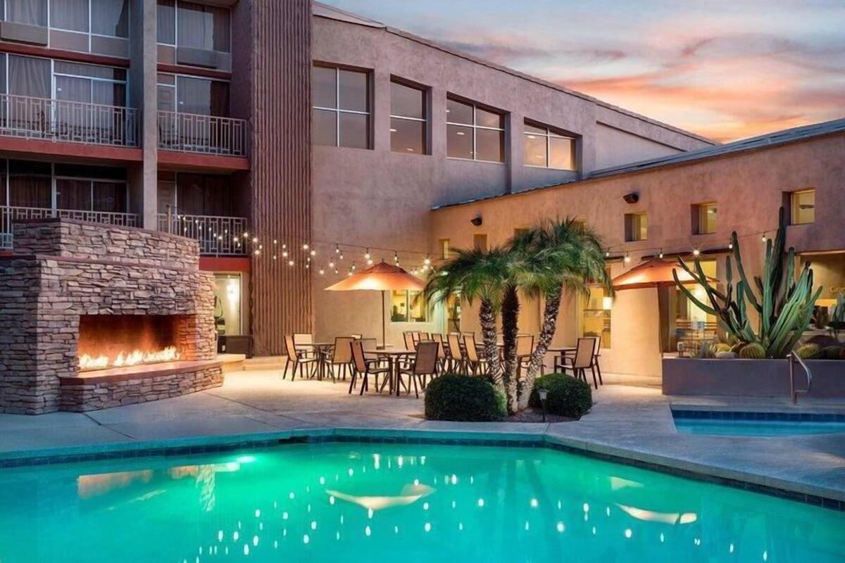 A hotel courtyard with a swimming pool, outdoor seating, a stone fireplace, and a building with balconies.