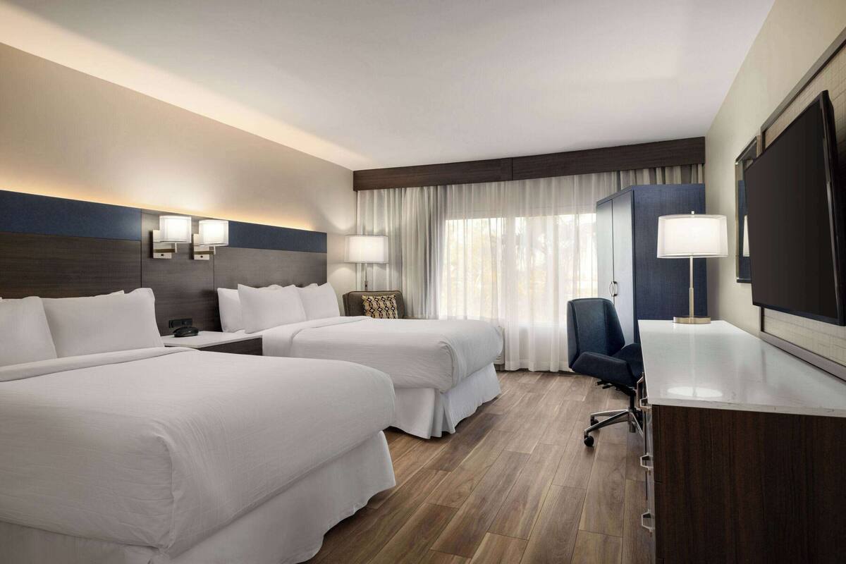 A hotel room with two beds, a desk, a chair, a TV, and a window with curtains.