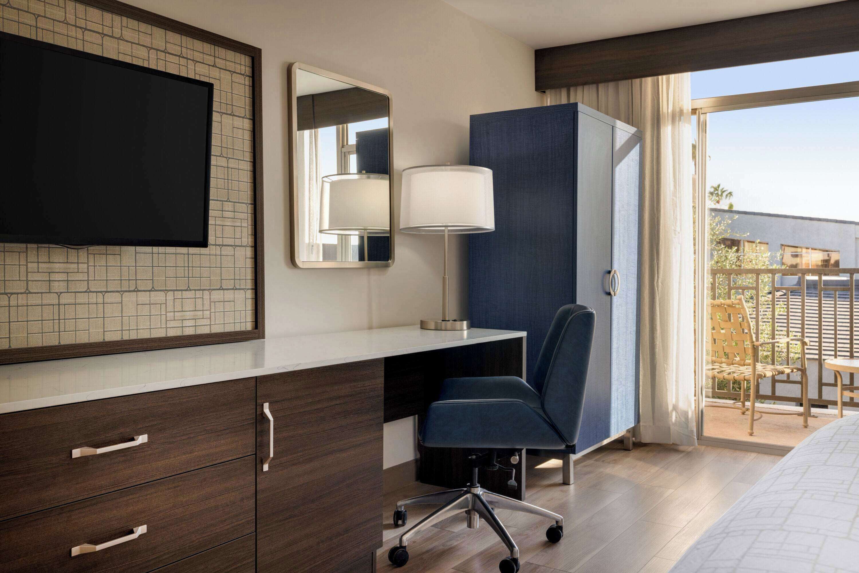 A hotel room with a flat-screen TV, a desk with a lamp, a chair, and a large window with a view of the outdoors.