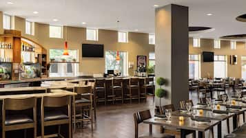A spacious restaurant with multiple dining tables and chairs, a bar area with shelves of bottles, and large windows allowing natural light.
