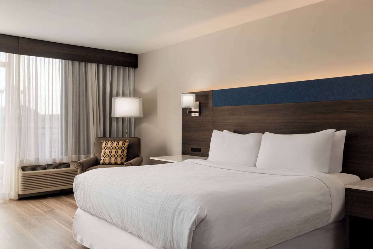 A hotel room with a large bed, a chair, a nightstand, a wall-mounted lamp, and a window with curtains.