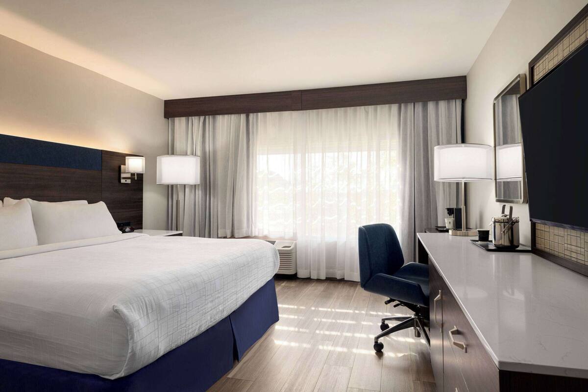 A modern hotel room with a large bed, a desk, a chair, and a large window with curtains.
